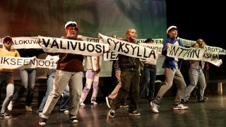 Musikaalista kuva, jossa tanssijoilla tekstejä sisältävät kyltit käsissä.